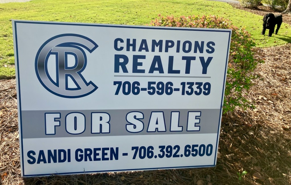 A corrugated yard sign displaying "For Sale" in bold letters, placed in front of a residential property.